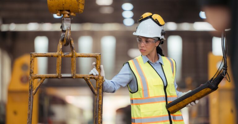 A woman stands near a crane. She has one hand on a part the crane is lifting and the other hand on the hoist control station.