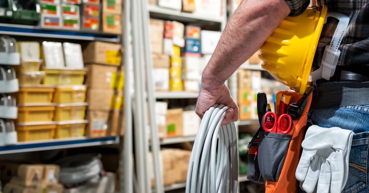 A man with a hard hat under his arm, a cable in his hand, gloves in his pocket, and a tool belt around his waist, in a storage room.