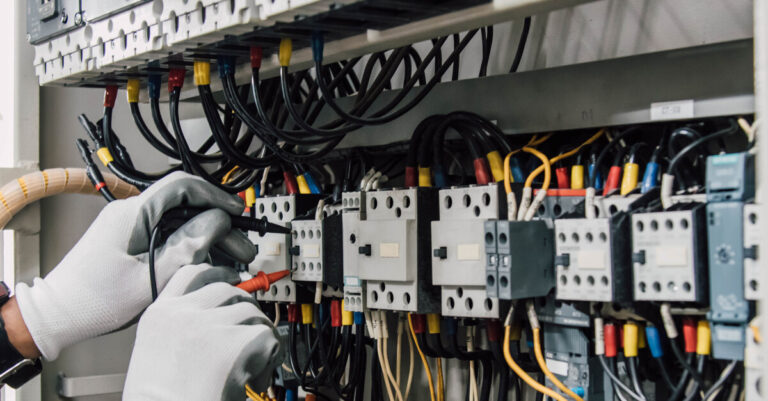 A pair of gloved hands connecting wires, one black, one red, to a relay protection system inside an electrical box.