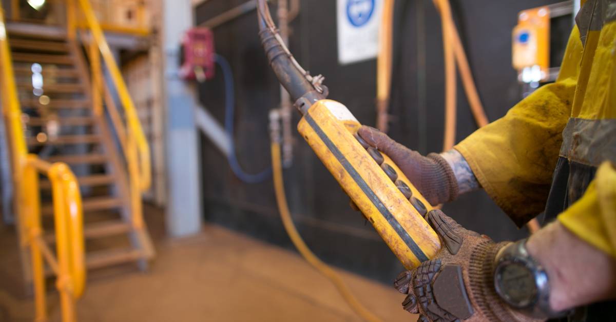 In an industrial facility, a person wearing a safety vest and gloves holds a pendant control station with both hands.