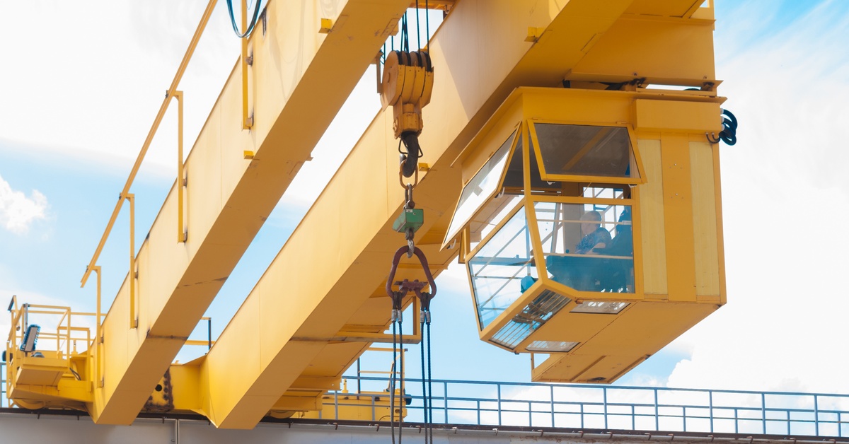 An operator sits in the glass cockpit of a yellow factory warehouse overhead hoist crane. He looks to the right.