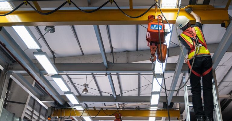 A worker in a high-visibility vest and hardhat inspects a crane hoist and hook while on a ladder in a factory.