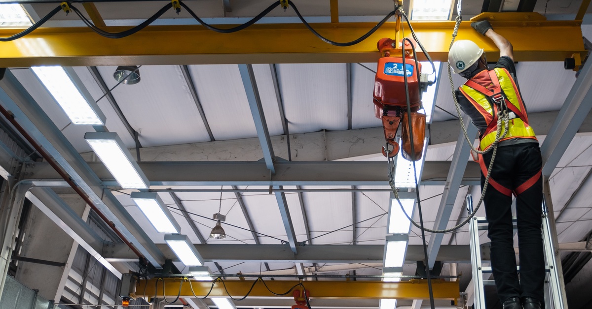 A worker in a high-visibility vest and hardhat inspects a crane hoist and hook while on a ladder in a factory.