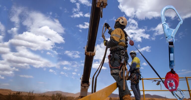 An industrial worker wearing a helmet and strapped to a harness stands on a raised platform to inspect the hook of a crane.