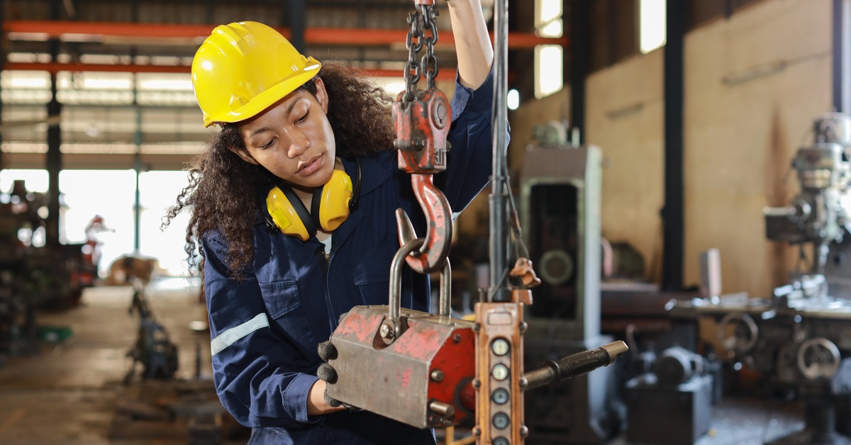 A woman in a yellow hard hat and a denim jumpsuit checks the control system of an overhead crane in an industrial warehouse.