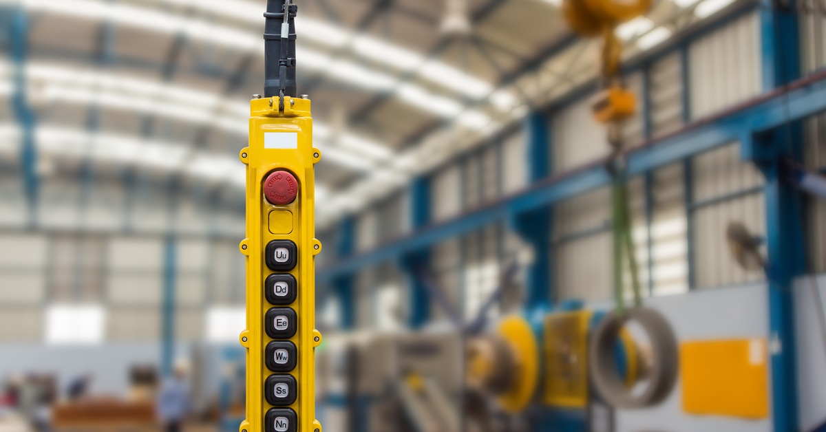 Yellow industrial pendant control station with push buttons and emergency stop switch hangs inside a factory workshop.