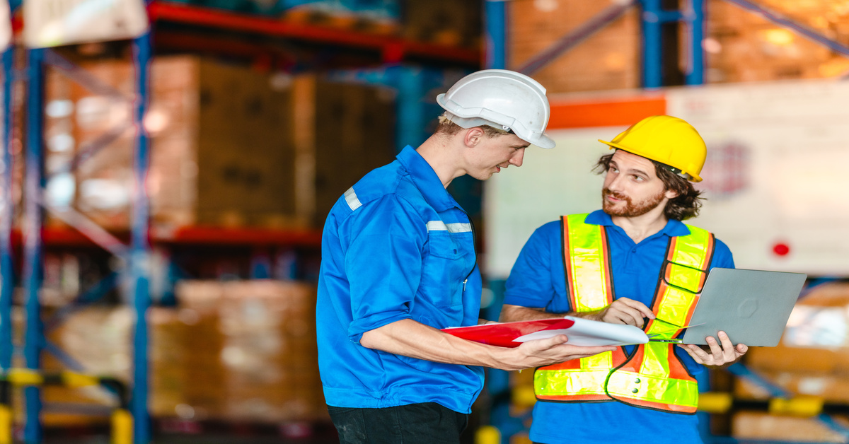 Two warehouse employees wearing hard hats review inventory information on a laptop and a clipboard.