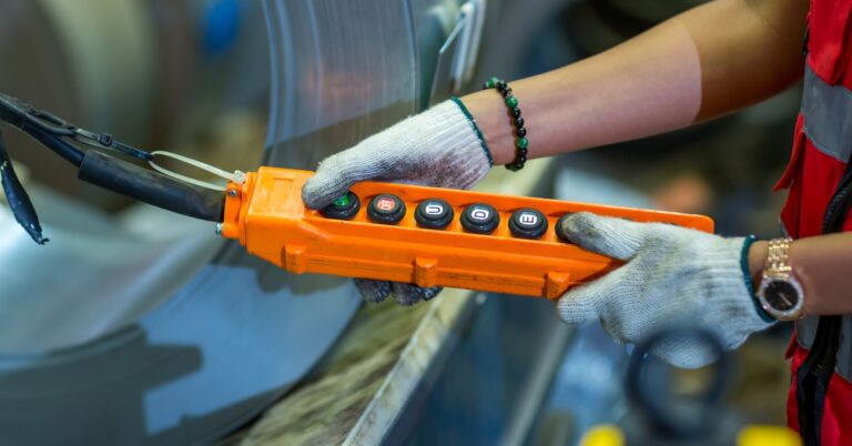 An industrial worker wears protective gloves as they operate an overhead crane using a pendant control station.