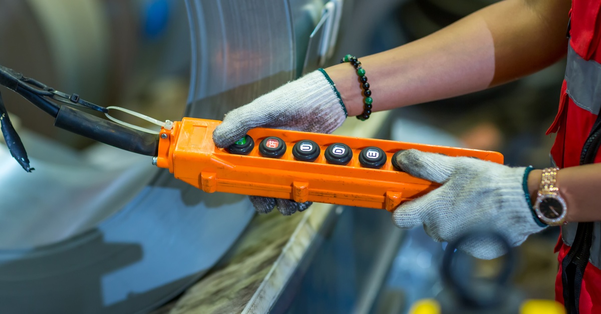 An industrial worker wears protective gloves as they operate an overhead crane using a pendant control station.
