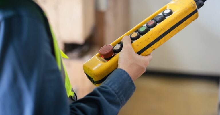 A worker holds a yellow crane pendant control with multiple buttons, operating lifting equipment in an industrial warehouse.