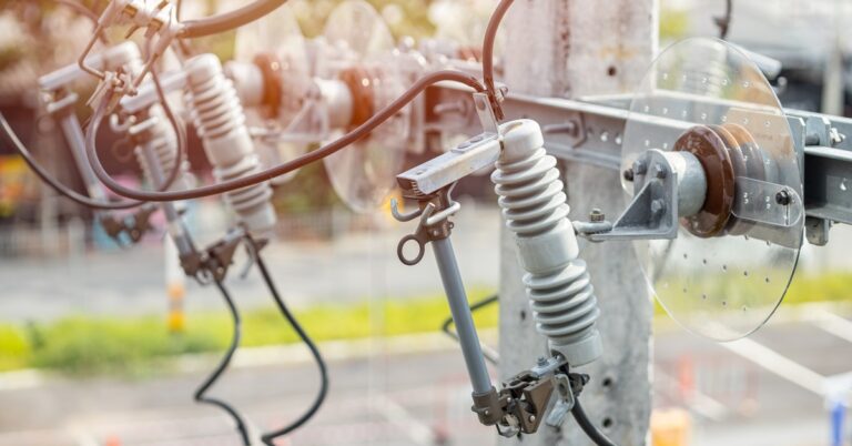 An electrical fuse cutout and insulators mounted on a utility pole with overhead power lines in an outdoor distribution system.