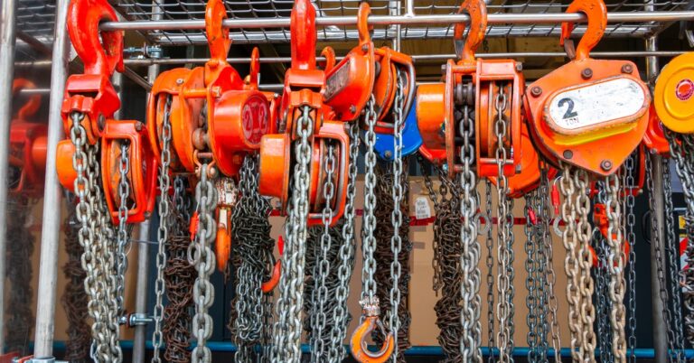 Multiple orange chain hoists with load chains hang on a metal storage rack, including a 2-ton unit, in an industrial setting.