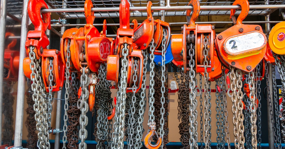 Multiple orange chain hoists with load chains hang on a metal storage rack, including a 2-ton unit, in an industrial setting.