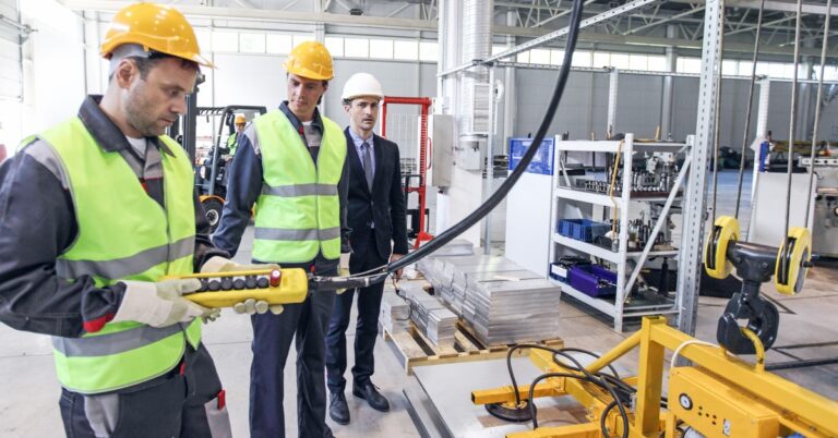 Three workers in hard hats operate a hoist controller on an industrial warehouse floor surrounded by heavy equipment.