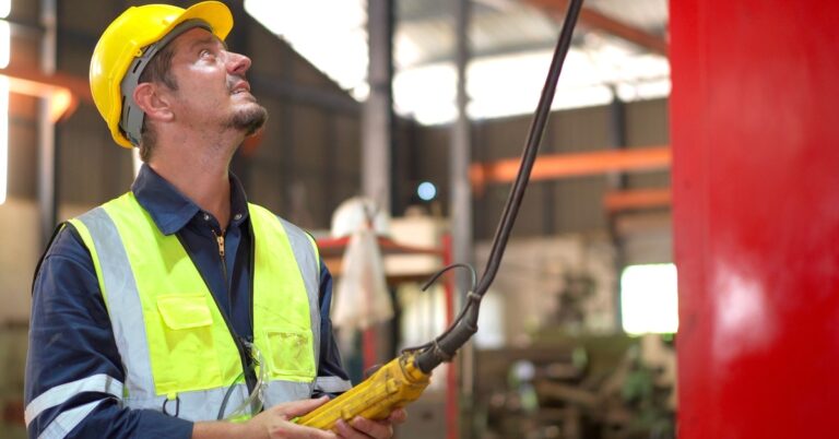 A worker in a safety vest and a hard hat operates a yellow pendant control station to direct an overhead crane in a warehouse.