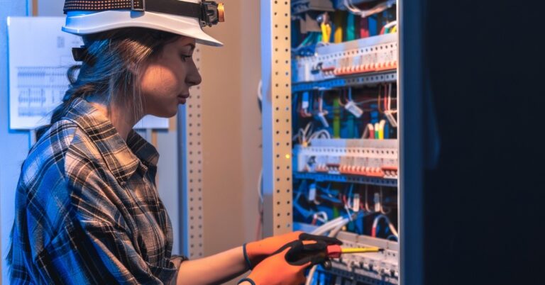 A woman in a flannel shirt, a hard hat, and work gloves repairs an electrical panel in a technical room.