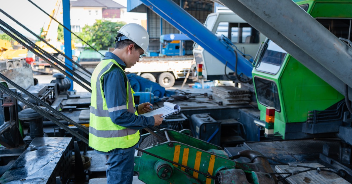A man in a hard hat and high-visibility vest looks at a clipboard on a mobile crane deck outside in front of a crane.