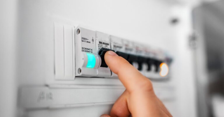 A person pushing a black switch on a fuse board upward. The switch sits next to a small, green light.